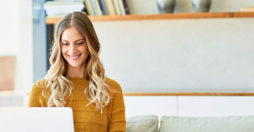 woman smiling at her laptop while sitting on the couch working remotely