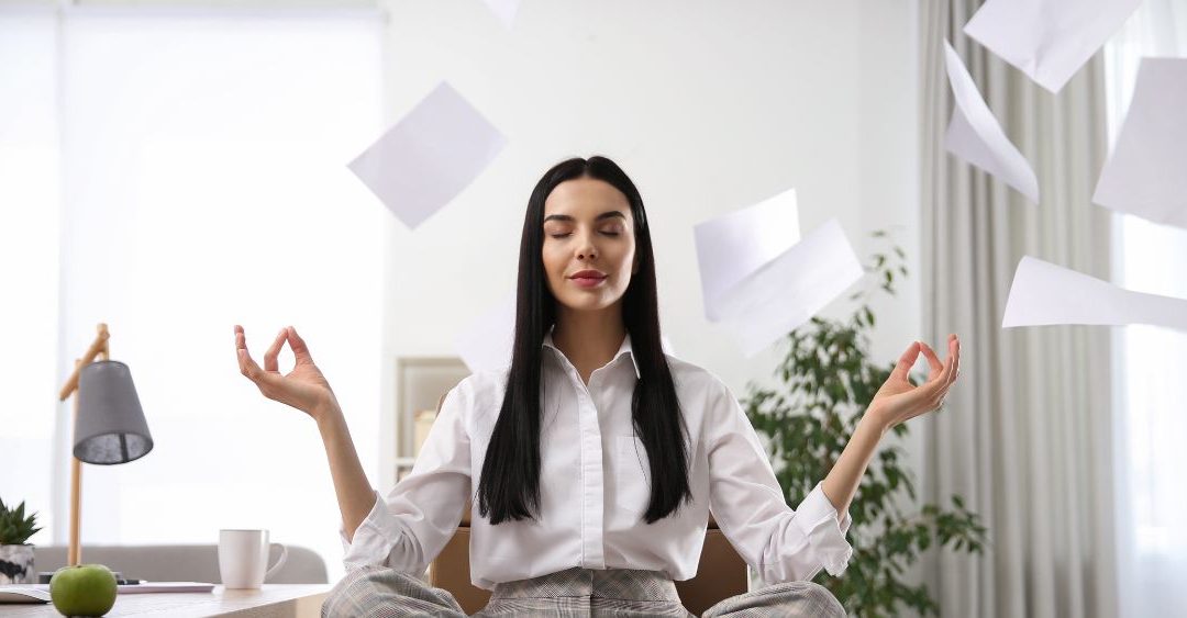 woman with long black hair and a white button up shirt and slacks in a meditation pose in her office with papers flying everywhere around her
