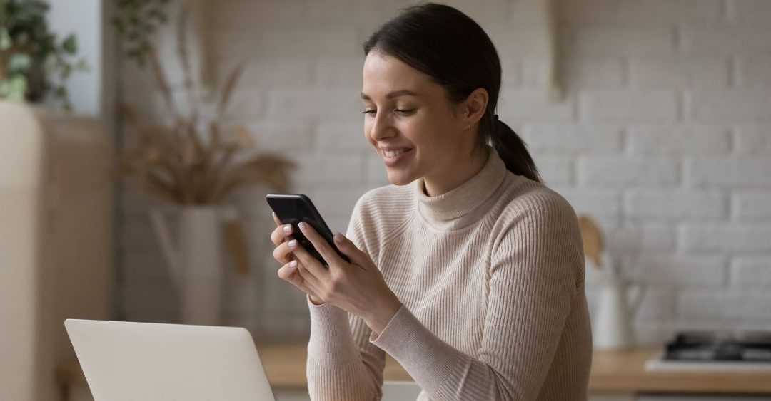 young white brunette with a cream sweater sitting in her home office looking at her phone while smiling